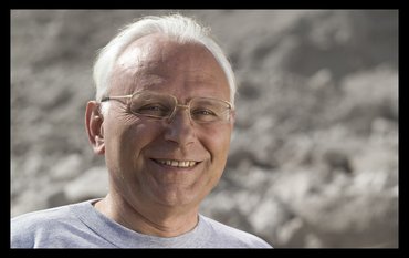 Porträtfoto eines Mannes mit grauen, kurzen Haaren, Brille und freundlichem Lächeln.