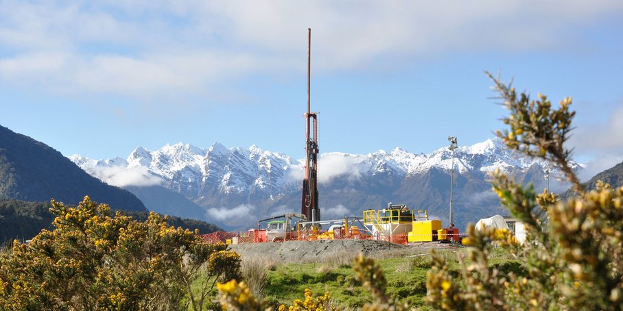 Mobile Bohranlage mit LKW und bunten Containern vor beschneiten Berggipfeln. Im Vordergrund: Blühende Büsche.