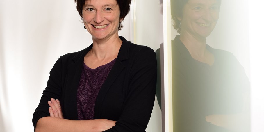 A portrait of a woman with short dark hair and a dark top. She is leaning against a glass wall with her arms crossed, smiling warmly, and her reflection is visible in the glass.