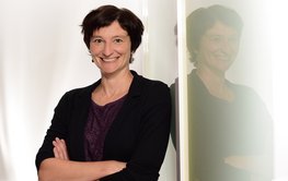 Porträt einer Frau mit dunklen kurzen Haaren und dunklem Oberteil. Sie lehnt freundlich lächelnd mit verschränkten Armen an einer Glaswand, in der sich ihr Bild spiegelt.