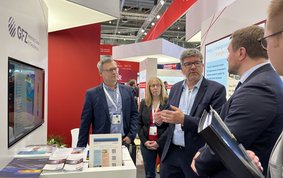 Fünf Personen stehen an einem Messestand mit einem weißen Tisch und einer Leinwand und unterhalten sich.