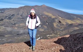 A young woman with long dark hair stands in a barren, mountainous landscape, wearing a white jacket, a cap and carrying a small rucksack.