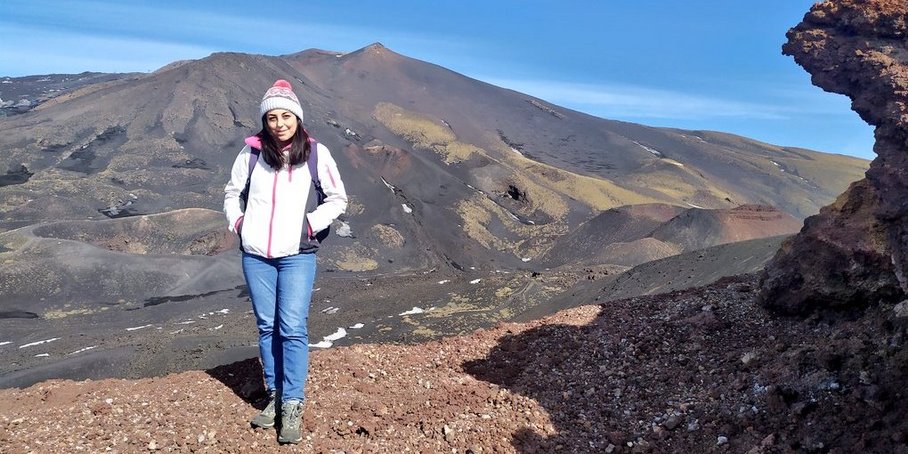 Eine junge Frau mit langen dunklen Haaren steht mit weißer Jacke, Mütze und mit kleinem Rucksack in einer kargen, bergigen Landschaft