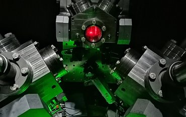 An assembly of metal components and flanges with thick bolts. The foreground is bathed in green light. Red light can be seen through a small round window.