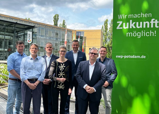 Group photo of seven people in front of a company building with a glass façade. ‘EWP’ is written above the entrance. To the right of the group is a grass-green wall bearing the slogan: ‘We make the future possible!’ ewp-potsdam.de