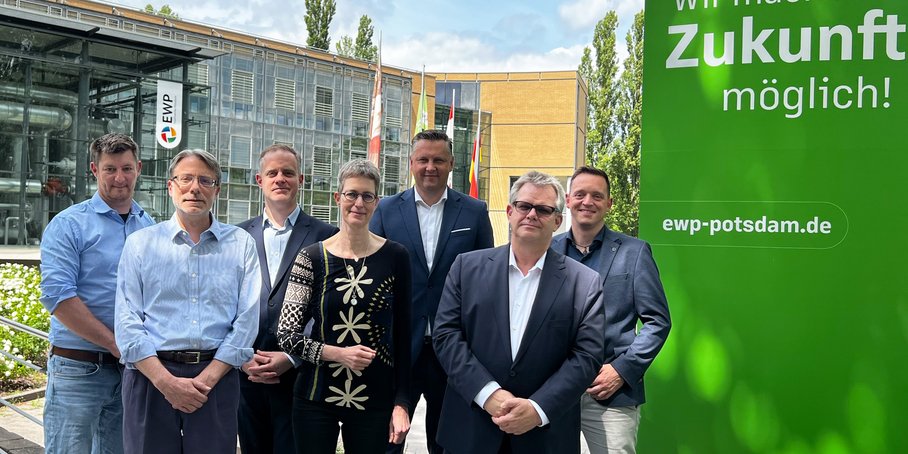 Group photo of seven people in front of a company building with a glass façade. ‘EWP’ is written above the entrance. To the right of the group is a grass-green wall bearing the slogan: ‘We make the future possible!’ ewp-potsdam.de