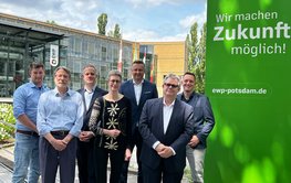 Group photo of seven people in front of a company building with a glass façade. ‘EWP’ is written above the entrance. To the right of the group is a grass-green wall bearing the slogan: ‘We make the future possible!’ ewp-potsdam.de
