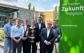 Gruppenfoto mit sieben Personen vor einem Firmengebäude mit Glasfront. Am Eingang steht EWP. Rechts neben der Gruppe ist eine grasgrüne Wand, auf der steht: Wir machen Zukunft möglich! ewp-potsdam.de