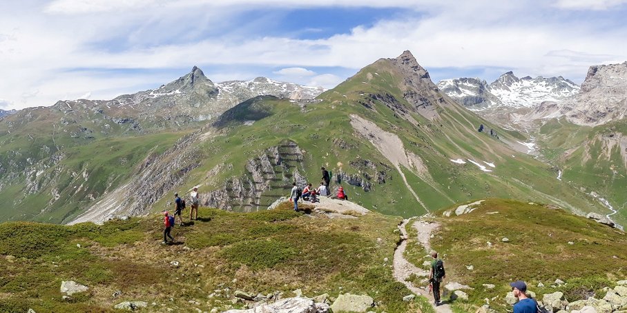 A typical Alpine landscape: jagged, rocky mountains, still covered in green vegetation in the foreground, and partly covered in snow in the background.
