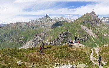 A typical Alpine landscape: jagged, rocky mountains, still covered in green vegetation in the foreground, and partly covered in snow in the background.