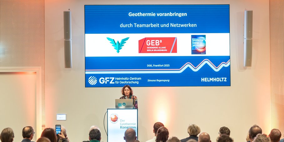 Eine Frau steht am Pult und hält einen Vortrag. Im Hintergrund auf großer Leinwand die Titelfolie der Präsentation: Text mit Logos. Im Vordergrund das Publikum.