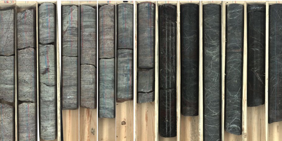 In einer unterteilten Holzkiste liegen 14 unterschiedliche lange, teils gebrochene grau-braune Bohrkerne nebeneinander. Die runden Stücke zeigen ein geschichtetes Muster.