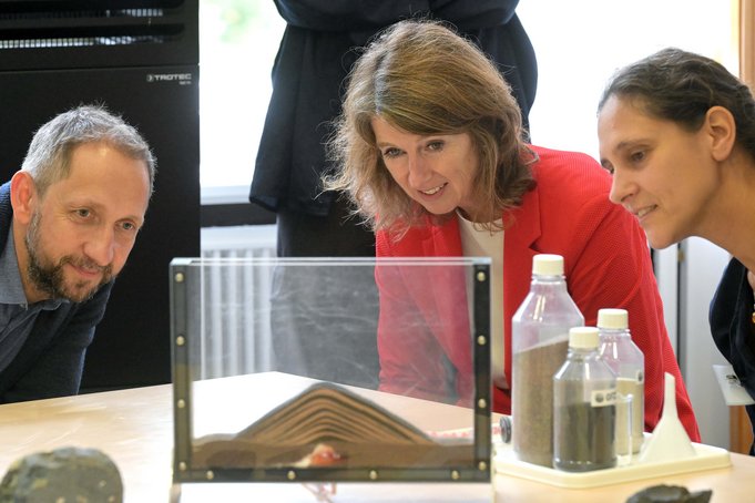 Drei Personen sitzen an einem Tisch vor einem Experiment: In einem flachen Glasaquarium ist verschiedenfarbiger Sand zu einem Berg aufgeschichtet.