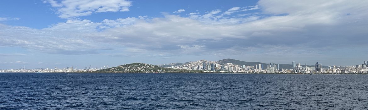 Vom Meer aus fotografiert: Eine riesige Küstenstadt auf Hügeln mit dichter Bebauung und vielen Hochhäusern. Im Vordergrund: Meer.