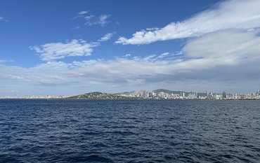 Vom Meer aus fotografiert: Eine riesige Küstenstadt auf Hügeln mit dichter Bebauung und vielen Hochhäusern. Im Vordergrund: Meer.