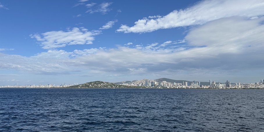 Vom Meer aus fotografiert: Eine riesige Küstenstadt auf Hügeln mit dichter Bebauung und vielen Hochhäusern. Im Vordergrund: Meer.