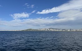 Vom Meer aus fotografiert: Eine riesige Küstenstadt auf Hügeln mit dichter Bebauung und vielen Hochhäusern. Im Vordergrund: Meer.