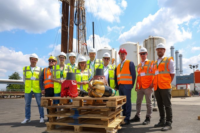 Gruppenfoto mit rund 10 Personen, die Warnwesten und Schutzhelm tragen. Sie stehen an einer Bohrung mit Bohrgestänge, das in den Himmel ragt. Im Hintergrund Türme eines Heizkraftwerks. Im Vordergrund auf Europaletten zwei Bohrmeißel.