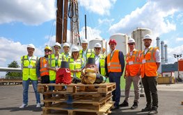 A group photo of around 10 people wearing high-visibility vests and hard hats. They are standing by a borehole with drill pipes stretching up into the sky. In the background are the towers of a combined heat and power plant. In the foreground, on Euro pallets, are two drill bits.