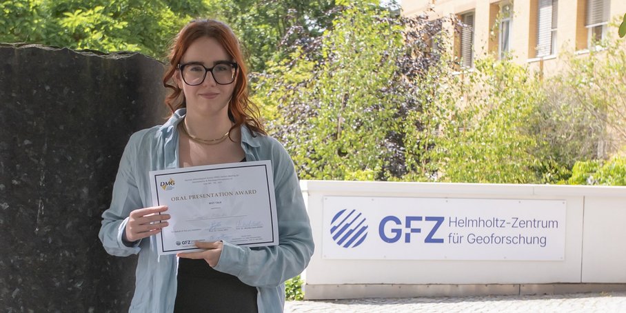 Auf dem Bild ist Candice Filoche zu sehen. Sie hält die Auszeichnung der DMG in ihren Händen. Im Hintergrund ist eine Säule des Säulenforums sowie das Logo des GFZ am Eingang zu Haus G zu sehen.