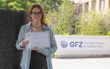 Auf dem Bild ist Candice Filoche zu sehen. Sie hält die Auszeichnung der DMG in ihren Händen. Im Hintergrund ist eine Säule des Säulenforums sowie das Logo des GFZ am Eingang zu Haus G zu sehen.