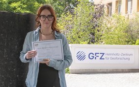 Auf dem Bild ist Candice Filoche zu sehen. Sie hält die Auszeichnung der DMG in ihren Händen. Im Hintergrund ist eine Säule des Säulenforums sowie das Logo des GFZ am Eingang zu Haus G zu sehen.
