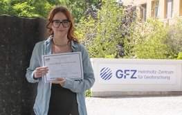 Auf dem Bild ist Candice Filoche zu sehen. Sie hält die Auszeichnung der DMG in ihren Händen. Im Hintergrund ist eine Säule des Säulenforums sowie das Logo des GFZ am Eingang zu Haus G zu sehen.