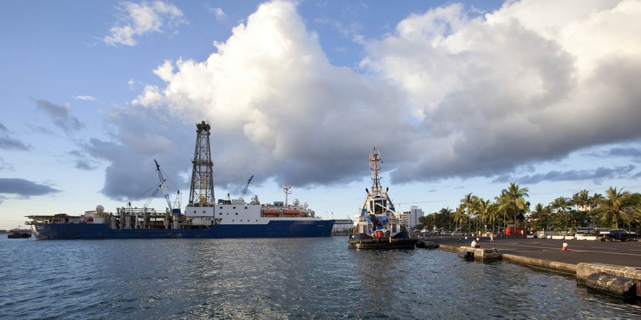 Das Forschungsschiff liegt im Hafen. Der Himmel ist bewölkt, aber blau. Das Meer ist ruhig. An Land ein asphaltierter Parkplatz mit Palmen direkt an der Hafenkante. Menschen sitzen und spazieren dort.