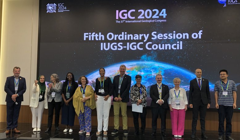 Gruppenbild mit 12 Personen, die nebeneinander vor einer Leinwand stehen. Die Projektion im Hintergrund zeigt einen Teil der Erde vor dunklem Hintergrund und das IUGS-Logo mit dem Titel der Veranstaltung.