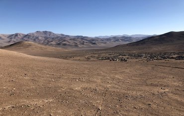 Brown, dry soil without plants with small stones. In the background: brown mountains, blue sky.