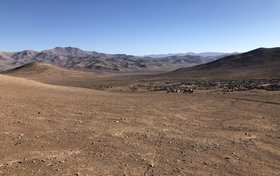 Brauner, trockener Boden ohne Pflanzen mit kleinen Steinen. Im Hintergrund: braune Berge, blauer Himmel.