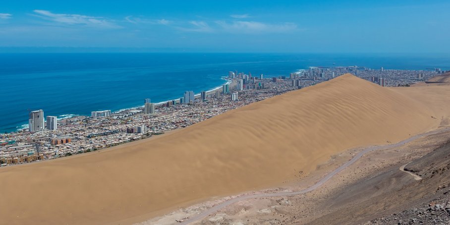 Landschaftsaufnahme: Im Vordergrund eine riesige Sanddüne, dahinter – wie ein Streifen – eine Küstenstadt am Meer. Im Hintergrund der blaue Ozean und blauer Himmel.
