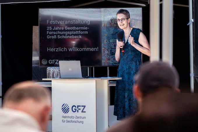 Eine Frau mit kurzem Haar und blauem Kleid steht vor einem Bildschirm, neben sich ein weißes Rednerpult. Im Vordergrund Köpfe der Zuhörenden.