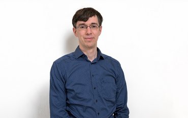 A middle-aged man wearing a blue shirt, glasses, and with short dark hair, standing in front of a white background.