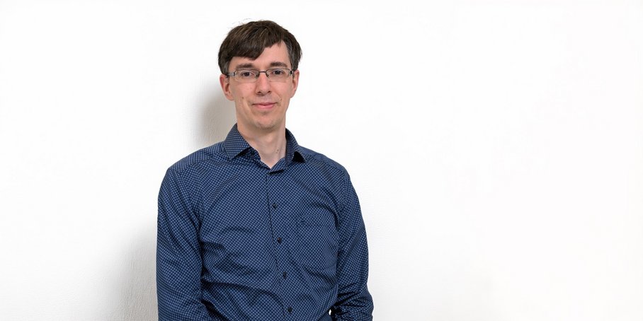 A middle-aged man wearing a blue shirt, glasses, and with short dark hair, standing in front of a white background.