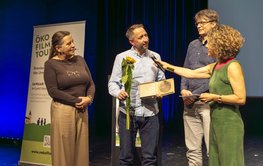 Vier Personen stehen auf einer Bühne, im Hintergrund links ein Rollbanner der Ökofilmtour 2026, während Blume und Urkunden an Martin Herold überreicht werden.