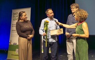 Vier Personen stehen auf einer Bühne, im Hintergrund links ein Rollbanner der Ökofilmtour 2026, während Blume und Urkunden an Martin Herold überreicht werden.