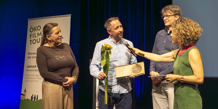 Vier Personen stehen auf einer Bühne, im Hintergrund links ein Rollbanner der Ökofilmtour 2026, während Blume und Urkunden an Martin Herold überreicht werden.