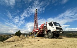 LKW mit ca 10 Meter hohem Bohrturm in einer kargen hügeligen Landschaft vor blauem Himmel mit Wolkenstreifen