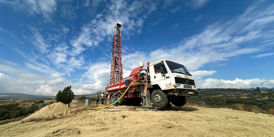 LKW mit ca 10 Meter hohem Bohrturm in einer kargen hügeligen Landschaft vor blauem Himmel mit Wolkenstreifen