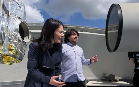 Two people are standing on the roof of a building among the sky. One is looking into a technical pipe.
