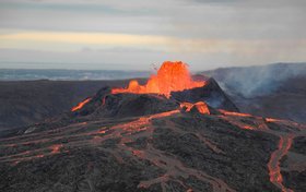 Dunkelgraue, flache Vulkanlandschaft, in der Mitte sprudelt rot-glühende Lava aus einem Krater und rinnt die Hänge entlang.