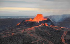 Dark grey, flat volcanic landscape, in the middle red-hot lava gushes out of a crater and runs down the slopes.