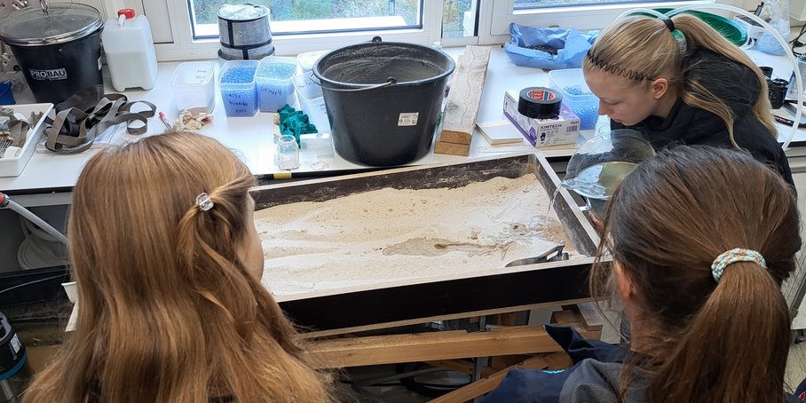 Three female students, photographed from behind, stand in front of a trough filled with sand that simulates river systems.