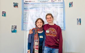 Two young adults stand arm in arm in front of a white wall with a scientific poster in the background. In front of them are two chair backs with jackets and a small glass column with different coloured sediment layers.