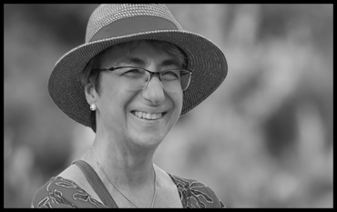 Black-and-white photograph: a portrait of a middle-aged woman. She is smiling and is wearing glasses, earrings and a hat.