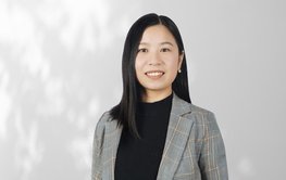 A portrait of a young woman with long black hair. She is wearing a black top and a grey checked blazer and is standing in front of a white wall on which the shadows of trees can be seen.