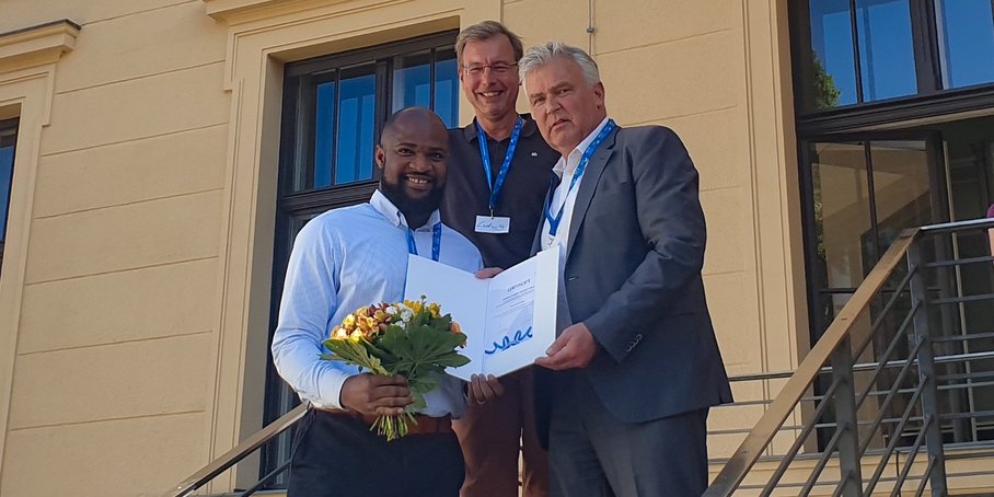 Three people are standing on a staircase in front of a yellow house: on the right, the award winner with a bouquet of flowers in his hand. He is being presented with the certificate.