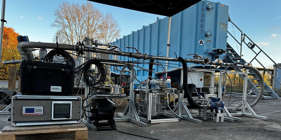 A multi-part measuring device comprising metal brackets, hoses, pipes and cables stands on a paved outdoor area next to a blue steel container, with a flight of steps leading up to it.