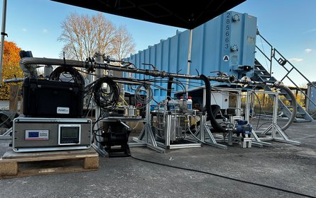 A multi-part measuring device comprising metal brackets, hoses, pipes and cables stands on a paved outdoor area next to a blue steel container, with a flight of steps leading up to it.
